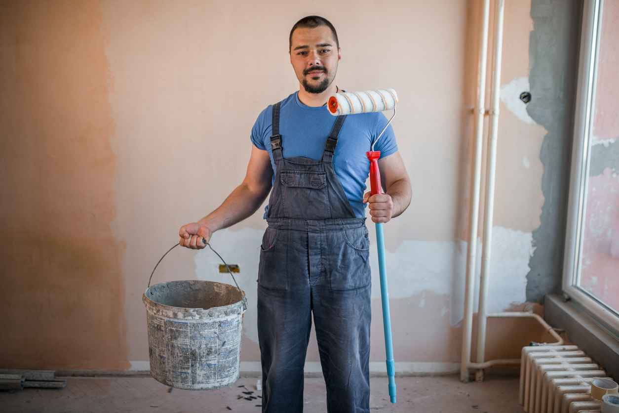 Smiling painter in coveralls holding paint roller and a bucket