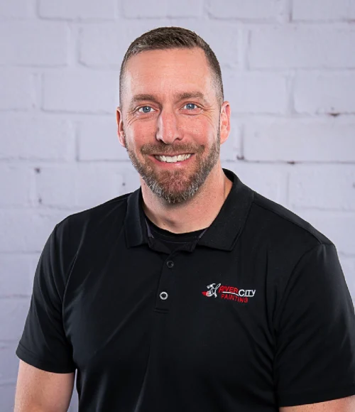 River City Painting Estimator Adam smiles for an updated professional headshot in 2025. He in his mid 30's and wears a black River City Painting embroidered polo shirt. Adam has blue eyes and short dark blonde hair and beard.