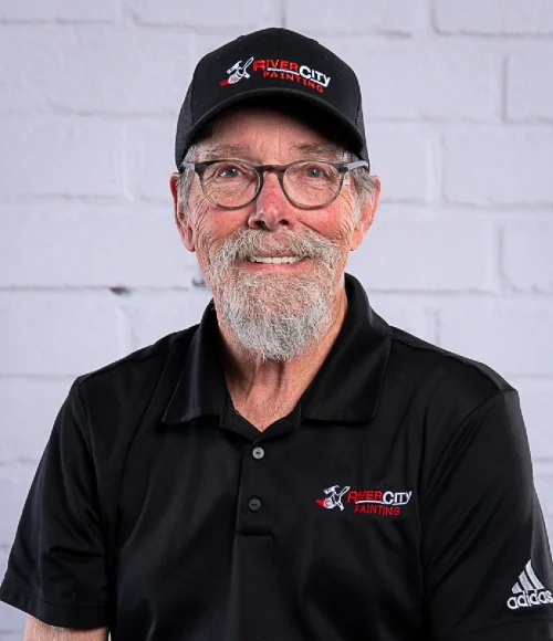 Project Manager, Jim Houser, poses for an updated professional portrait in 2025. Jim is wearing a black River City Painting embroidered baseball cap and black button up polo shirt. Jim is in his 60's with blue eyes and grey hair/beard. He has a nice smile and wears glasses.